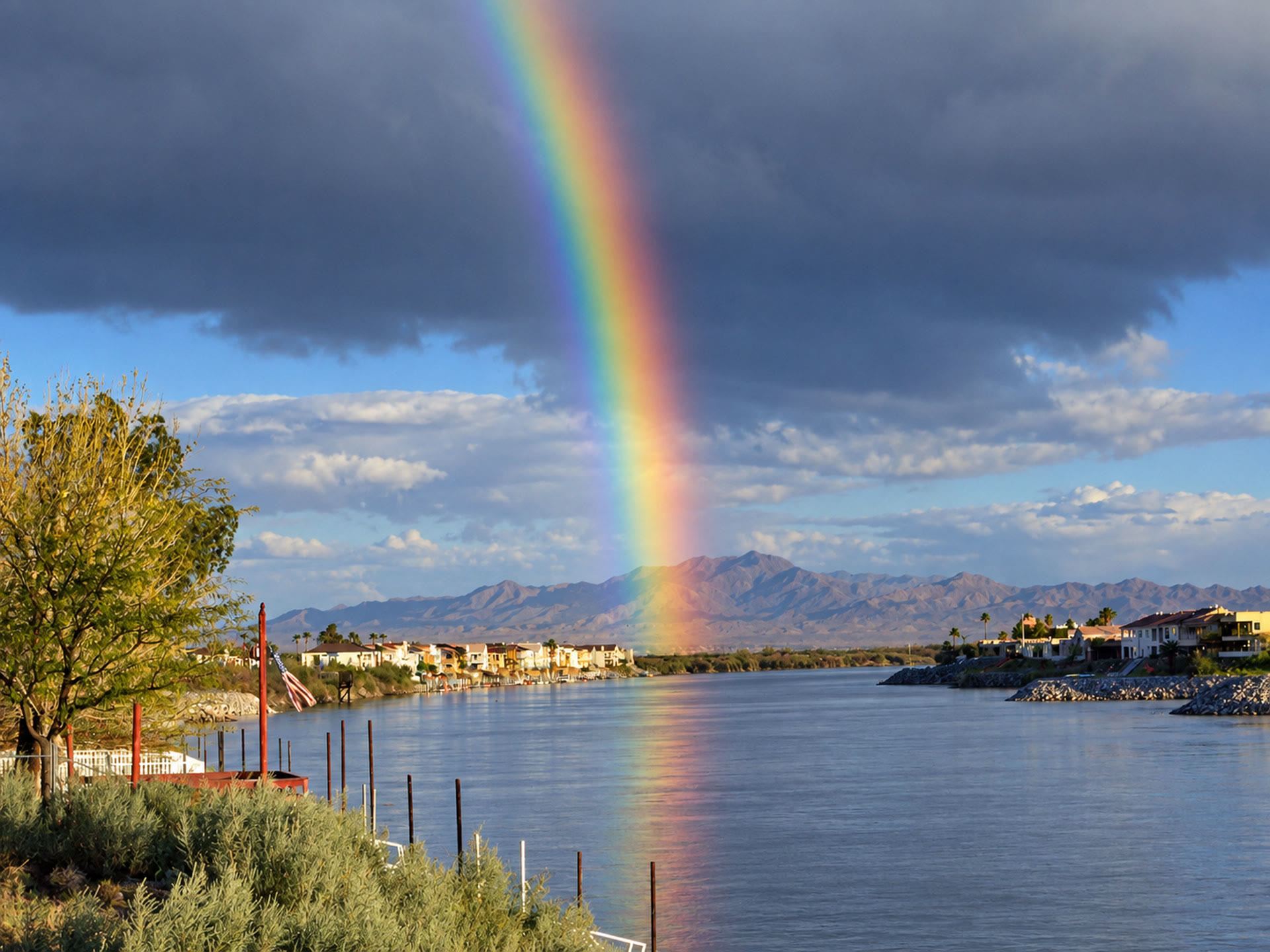 Bright rainbow reflecting on the Colorado River, with waterfront homes, desert mountains and storm clouds in the background.