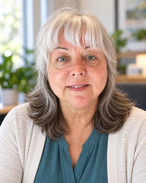 Barbara "Bobbie" McClure wearing a teal blouse and cream cardigan, with a bright window and plants behind her.